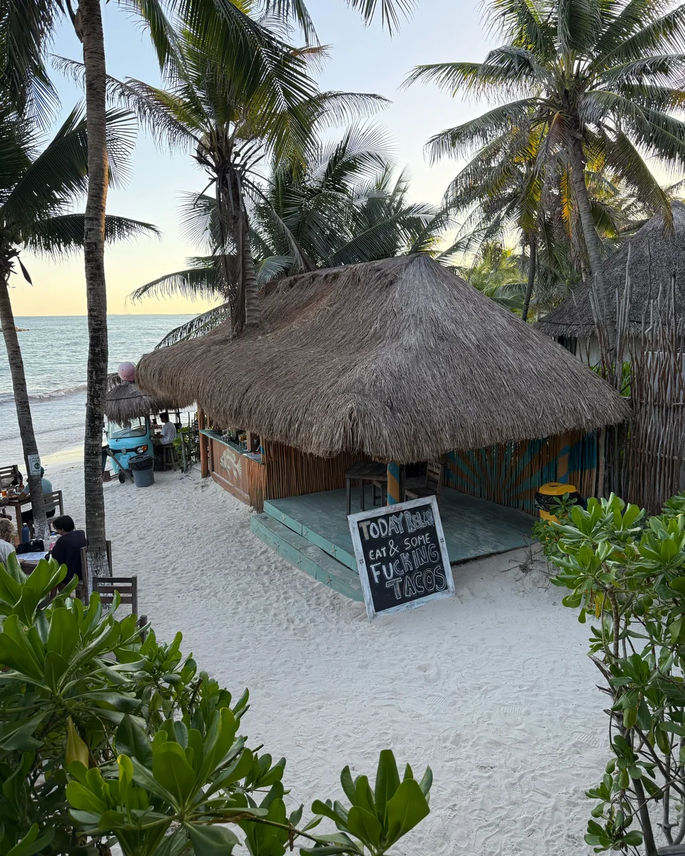 Una palapa de techo de palma sobre una playa de arena blanca entre palmeras, con el mar al fondo y un letrero de pizarra en la arena.