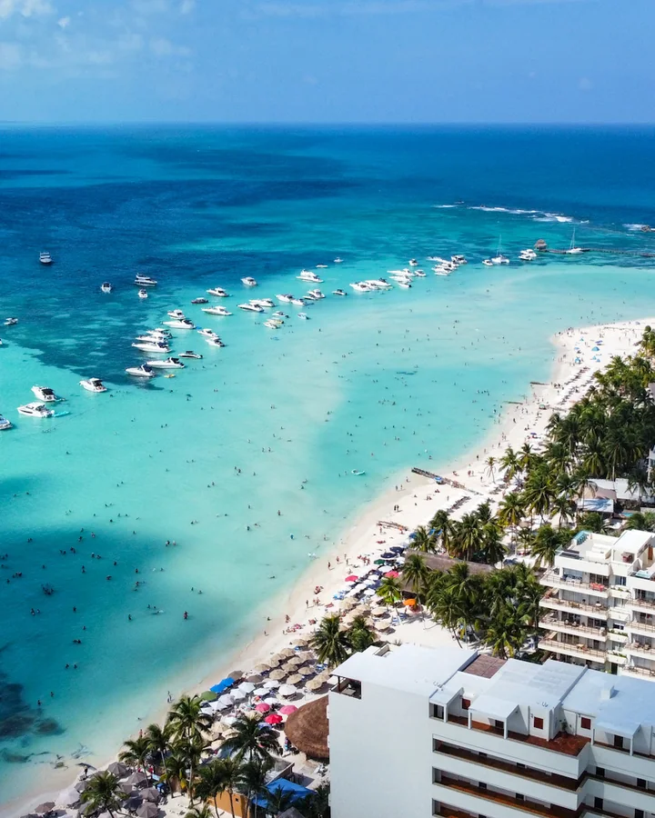 Vista aérea de aguas turquesa poco profundas con numerosas embarcaciones, una playa de arena blanca que se curva junto a una orilla de palmeras y edificios, y un mar azul intenso hasta el horizonte.