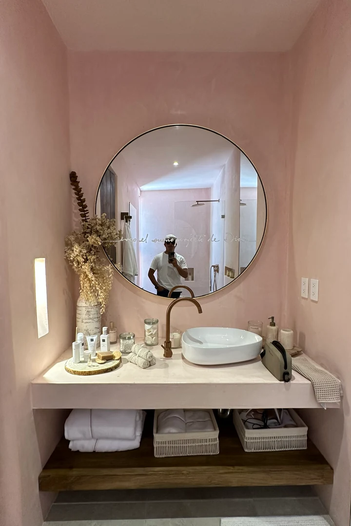 Lavabo en tonos rosa con espejo circular, grifería de cobre y flores secas, con la persona que toma la foto reflejada en el espejo, durante una estancia en Tulum.