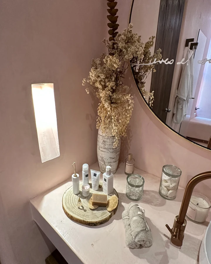 Detalle de un lavabo rosa: flores secas en un jarrón, artículos de baño sobre una bandeja de madera, una toalla enrollada, frascos de vidrio y una grifería de cobre junto a un nicho de pared con luz suave.